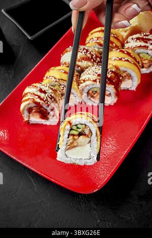 Nahaufnahme der weiblichen Hand mit Stäbchen zum Aufnehmen appetitlicher Sushi-Brötchen gefüllt mit Aal, Gurke und Frischkäse mit Cheddar von VIBRANT Stockfoto