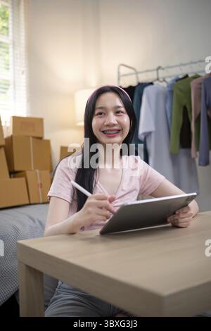 Glückliche junge Frau mit Zahnspangen, die selbstbewusst ihren Online-Shop verwaltet und ein Tablet in ihrem einladenden Arbeitsplatz in der Hand hält, umgeben von Paketen Stockfoto