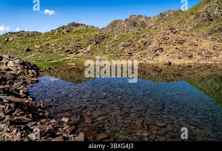 Eine ruhige Berglandschaft mit einem klaren See, der felsige Hügel und blauen Himmel reflektiert. Im Vordergrund stehen dunkle Steine und üppiges grünes Gras, Crea Stockfoto
