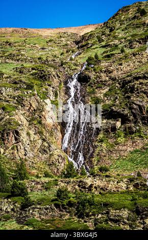 Ein atemberaubender Wasserfall, der einen felsigen Berghang hinunterstürzt, umgeben von üppigem Grün und rauem Gelände unter einem klaren blauen Himmel. Stockfoto