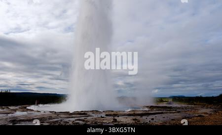 Strokkur Geysir bricht gewaltig in die Luft aus, umgeben von einer zerklüfteten Landschaft in Island. Besucher bestaunen dieses Naturwunder, das die Schönheit der geothermischen Aktivität in der Region zeigt. Stockfoto
