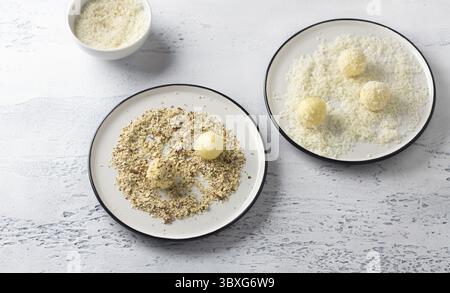 Essen, weiße Schokoladentrüffel in Kokosflocken gerollt und zerstoßene Nüsse auf einem weißen Teller, Blick von oben. Kochstufe mit köstlichen Pralinen Stockfoto