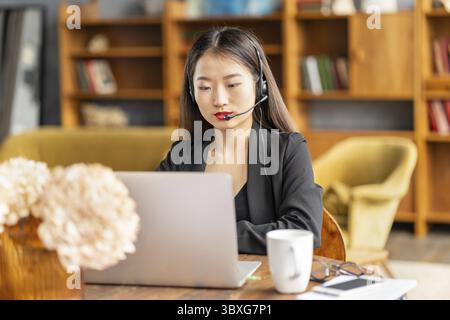 Asiatische Geschäftsfrau im Headset spricht per Konferenzschaltung und Video-Chat auf Laptop im Büro, lächelnd chinesische Support-Service-Agent-Beratung individuell Stockfoto