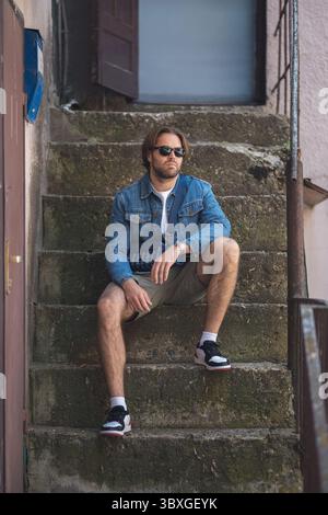 Ein junger Mann in Jeansjacke und weißem T-Shirt sitzt auf der Außentreppe und lächelt. Legere Sommermode mit Sonnenbrille, Shorts und Sneakers. Glücklich, rel Stockfoto