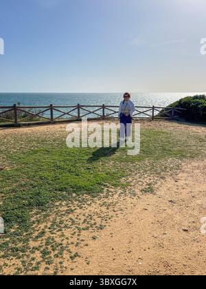 Frau in der Mitte des Lebens steht auf Gras in der Nähe des Ozeans, gekleidet in legere Kleidung, umgeben von natürlicher Schönheit, verkörpert Wellness und friedliches Leben Stockfoto
