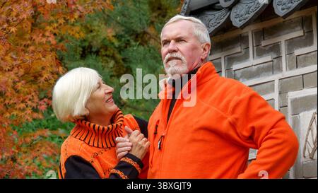 Älteres grauhaariges optimistisches Ehepaar Mann und Frau vor dem Hintergrund eines japanischen Pavillons im Herbst in orangefarbenen Pullis Stockfoto