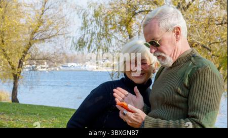 Porträt eines älteren grauhaarigen Paares, eines Mannes in Khaki-Militärkleidung, einer Frau in einem schwarzen Pullover, mit einem orangen Smartphone Stockfoto