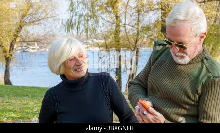 Porträt eines älteren grauhaarigen Paares, eines Mannes in Khaki-Militärkleidung, einer Frau in einem schwarzen Pullover, mit einem orangen Smartphone Stockfoto