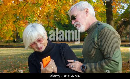 Porträt eines älteren grauhaarigen Paares, eines Mannes in Khaki-Militärkleidung, einer Frau in einem schwarzen Pullover, mit einem orangen Smartphone Stockfoto