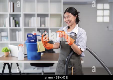 Lächelnde Büroreinigerin, die einen Staubsauger hält und ein Telefon benutzt, wodurch das Multitasking am Arbeitsplatz hervorgehoben wird Stockfoto