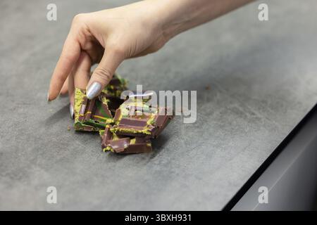 Die Hand der Frau hält ein Stück Dubai-Schokolade auf grauem Hintergrund. Grüner Kadayif und Pistazien mit Pistazien im Trenddessert. Nahaufnahme, selektiv Stockfoto