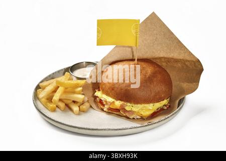 Köstlicher Burger in weißem Weizenbrötchen mit Hähnchenpastete, gebratenen Speckscheiben, Tomaten und Käsesauce, in Papier gewickelt, serviert auf Keramikplatte mit Stockfoto