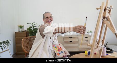 Ältere Frau, die zu Hause malte, lächelnd und mit dem Pinsel hielt, sich mit kreativer Kunst beschäftigte, helle und fröhliche Atmosphäre Stockfoto