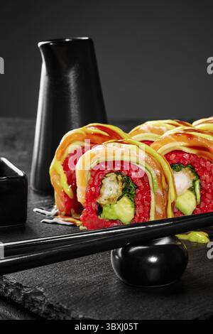 Frisch zubereitete Uamaki-Sushi-Brötchen mit Lachs, Avocado und lebendigem rotem Reis, serviert auf einem eleganten schwarzen Schieferteller mit Essstäbchen Stockfoto
