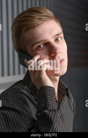 Ein junger Mann in einem schwarzen Hemd, der auf dem Handy auf dunklem Hintergrund spricht Stockfoto
