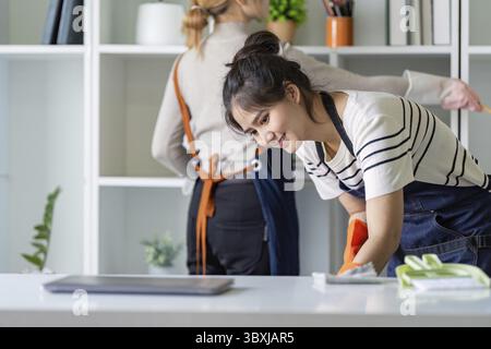 Junge Frau mit Schürze, lächelt beim Abstauben von Regalen in einem Büro und betont die Sauberkeit Stockfoto