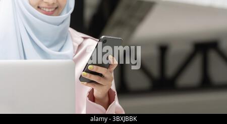 Eine Nahaufnahme einer muslimischen Frau im Hijab, die lächelt, während sie ein Smartphone hält, was eine effektive Kommunikation im modernen Geschäft symbolisiert Stockfoto