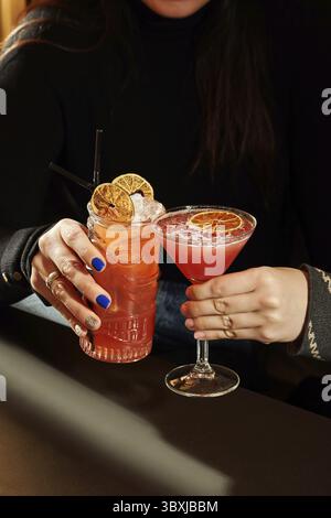 Frau genießt zwei erfrischende Cocktails, garniert mit getrockneten Orangen, einer in einem Tiki-Becher und der andere in einem Martini-Glas Stockfoto
