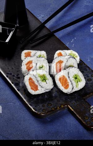Frisch zubereitete Uamaki-Sushi-Brötchen, gefüllt mit Lachs und Gurke, serviert auf einem dunklen Servierbrett mit Sojasauce und Essstäbchen, bereit zum en Stockfoto