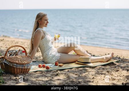 Romantische blonde Frau mit Strohhut beim Picknick am Strand. Frisches Obst, Saft, Kekse und Pfirsiche. Stockfoto