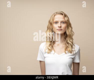 Porträt der schönen jungen, ernsten, klugen blonden Frau ohne Make-up an beigefarbener Wand. Hübsche Frau mit lockigen Haaren in weißem T-Shirt Stockfoto