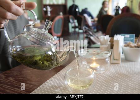 Frau, die grünen Tee aus der Teekanne aus Glas in Tassen im Café mit verschwommenem Hintergrund gießt. Frau, die grünen Tee aus der Teekanne in Tassen im Café mit gießt Stockfoto