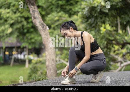 Eine junge Frau in sportlicher Kleidung bindet sich ihre Schnürsenkel in einem üppig grünen Park, macht sich bereit für Outdoor-Sport oder Joggen Stockfoto