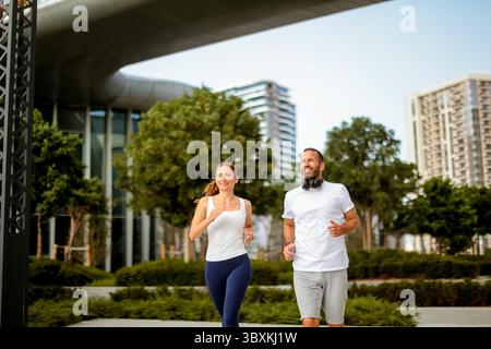 Beim Joggen in einem lebhaften Stadtpark strahlen ein Mann und eine Frau Energie und Gesellschaft aus. Umgeben von moderner Architektur, umarmen sie ein gesundes A Stockfoto
