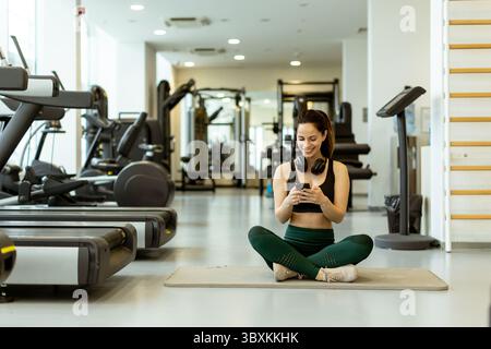 Lächelnde Frau sitzt im Kreuz auf einer Yogamatte mit Kopfhörern und konzentriert sich auf ihr Telefon in einer lebendigen, gut ausgestatteten Fitnessumgebung. Stockfoto