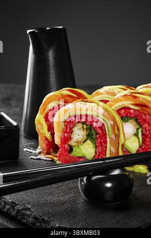Frisch zubereitete Uamaki-Sushi-Brötchen mit Lachs, Avocado und lebendigem rotem Reis, serviert auf einem eleganten schwarzen Schieferteller mit Essstäbchen Stockfoto