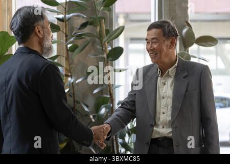 Ein fröhliches Treffen zwischen zwei älteren Männern, bei dem Respekt und Kameradschaft zum Ausdruck kommen, während sie sich gegenseitig mit einem Handschlag in einer stilvollen Umgebung begrüßen Stockfoto