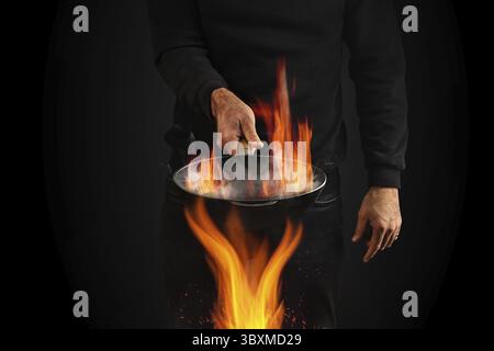 Junger Mann in Pullover und Jeans gekleidet. Er hält brennende Wok Pfanne mit Rauch über Feuer, vor schwarzem Studio Hintergrund. Kochkonzept. Nahaufnahme Stockfoto