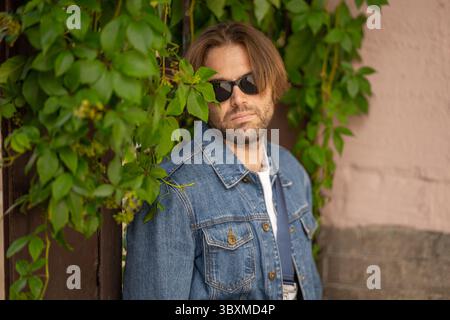 Ein stilvoller junger Mann in Jeansjacke und Sonnenbrille posiert vor einer rustikalen Wand mit wilden Trauben. Nahaufnahme. Stockfoto