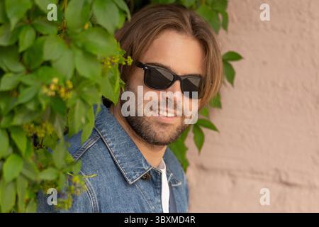 Ein stilvoller junger Mann in Jeansjacke und Sonnenbrille posiert vor einer rustikalen Wand mit wilden Trauben. Nahaufnahme. Stockfoto