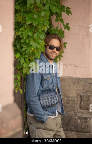 Ein stilvoller junger Mann in Jeansjacke und Sonnenbrille posiert selbstbewusst vor einer rustikalen Wand, die mit wilden Trauben bedeckt ist und coole urbane Vibes ausstrahlt. Stockfoto