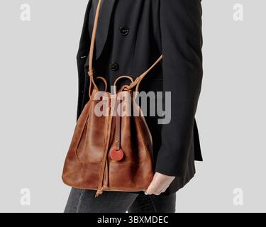 Nahaufnahme einer stylischen Frau mit lässiger schwarzer Jacke und Jeans, die eine schicke braune Ledertasche mit Kordelzug über der Schulter vor neutralem Hintergrund hält Stockfoto