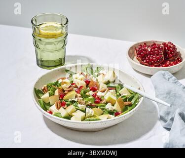 Obstsalat mit Nüssen, ausgewogenes Essen, sauberes Essen. Spinat mit Äpfeln, Pekannüssen und Feta, garniert mit Granatapfelkernen in einer Schüssel auf dem Tisch mit Weißwein Stockfoto
