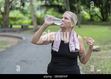 Eine ältere Frau mit Tattoos trinkt nach ihrem Training in einem malerischen Park Wasser und unterstreicht ihr Engagement für Fitness und Wellness Stockfoto