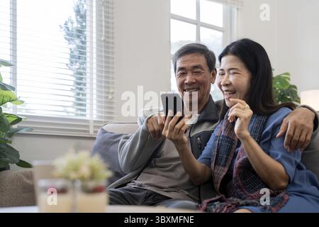 Glückliches älteres asiatisches Paar, das Smartphone benutzt, sitzt auf dem Sofa und macht E-Commerce-Shopping online auf der Website zu Hause Stockfoto