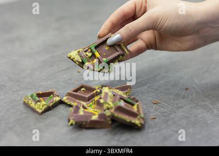 Die Hand der Frau hält ein Stück Dubai-Schokolade auf grauem Hintergrund. Grüner Kadayif und Pistazien mit Pistazien im Trenddessert. Nahaufnahme, selektiv Stockfoto