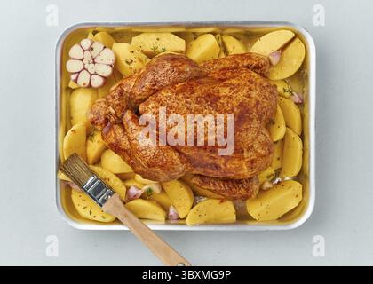 Ganzes mariniertes Hühnchen in Soße mit rohen Kartoffeln, mit Bürste in Backform, Blick von oben Stockfoto