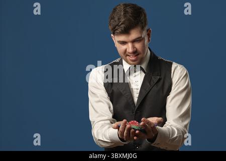 Gutaussehender Mann, Neuling im Poker, in schwarzer Weste und weißem Hemd. Halten Sie einige farbige Chips, lächeln Sie und schauen Sie sich sie an. Posiert gegen den blauen Studiobackgr Stockfoto