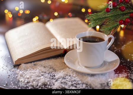 Heißer Tee, heiße Schokolade, Kaffee in einer weißen Tasse. Ein altes offenes Buch und ein Zweig des Neujahrsbaums. Verschwommenes Licht, dunkler hölzerner Hintergrund in weißem Schnee Stockfoto