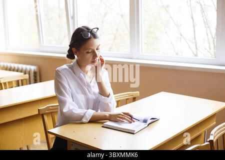Kaukasische Studentin sitzt an Schreibtischen, macht Notizen an der Universität, denkt und schreibt in einem Notizbuch an die Lehrerin, die einen Vortrag in einem College hält Stockfoto