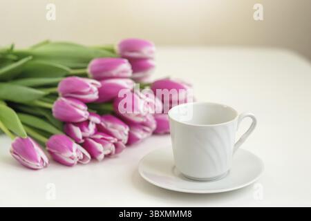 Ein weißer Becher und ein Blumenstrauß aus wunderschönen rosa Tulpen liegen auf einem weißen Tisch. Nahaufnahme. Kopierbereich Stockfoto
