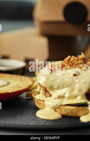 Nahaufnahme eines offenen Burgers mit paniertem Patty in cremiger Käsesauce, bestreut mit knusprigen gebratenen Zwiebeln, mit geröstetem Topbrötchen an der Seite, s Stockfoto