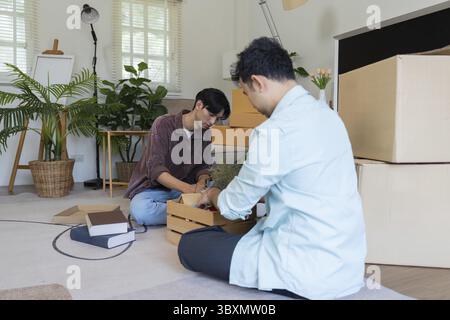 Zwei Männer, ein Paar, packten gerne Kisten in ihrem wunderschön eingerichteten Wohnzimmer aus, gefüllt mit Pflanzen und modernen Möbeln Stockfoto