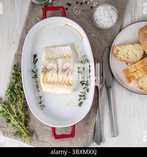 Kabeljaufilet, frisch im Ofen mit Thymian in einer Porzellanform zum Backen gekocht, Brot auf einem Teller, Salz und Pfeffer, weißer Holztisch, Blick von oben Stockfoto