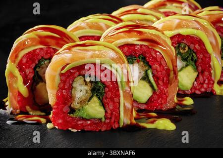 Makroaufnahme dekadenter Sushi-Brötchen mit Garnelentempura, bedeckt mit Lachsscheiben und mit herzhaften Saucen getränkt, serviert auf schwarzer Steinoberfläche für Stockfoto