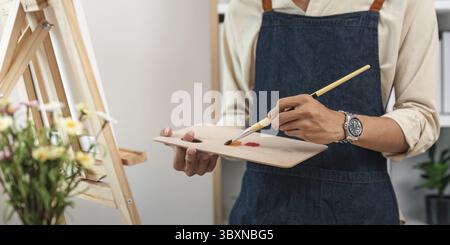 Nahaufnahme eines Künstlers auf Leinwand in einem Atelier, der eine Denim-Schürze trägt und eine Palette mit verschiedenen Farben hält Stockfoto